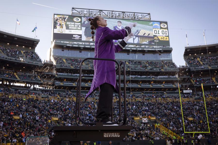 A student stand on an elevated platform and conducts with a Lambeau Field sign behind them.